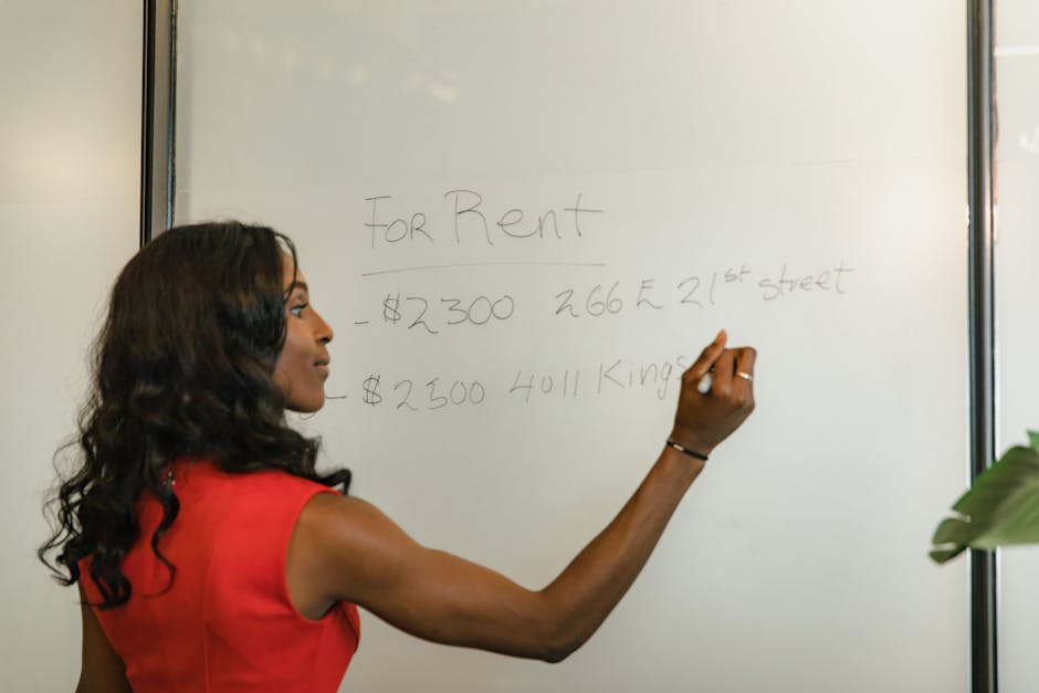 A woman with long, dark, wavy hair wearing a sleeveless red dress is standing in front of a whiteboard, writing with a black marker. The whiteboard has handwritten text indicating rental prices and addresses, with the words 'For Rent' at the top, along with figures and address details. The environment appears to be an office or meeting room with a bright, well-lit atmosphere, and part of a green plant is visible on the right side of the frame. The scene reflects an indoor setting used for discussions or presentations, with no visible rubbish or waste management elements. This image is relevant to scenarios involving professional communication or planning within a property or business context, subtly aligning with the idea of independent or private arrangements, akin to alternative waste handling or on-site clearance discussions, relevant to waste management service discussions at Rubbish Clearance Stockwell.