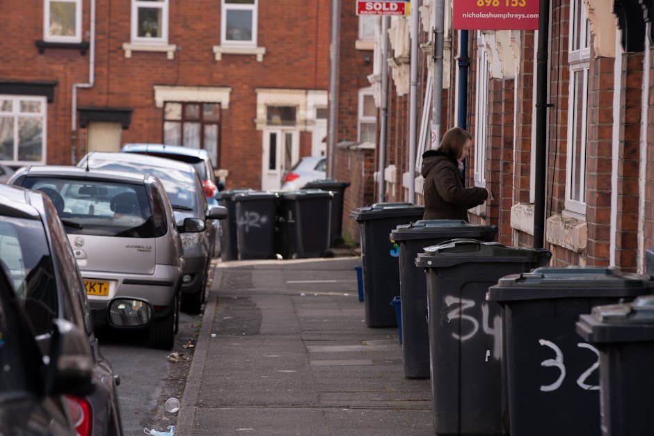 The image shows a residential street with a row of parked cars on the left side, including a silver car and a grey car directly adjacent to the grey concrete sidewalk. Multiple dark grey wheelie bins with closed lids are lined up along the right side of the sidewalk, each marked with white graffiti or spray-painted numbers, and positioned next to brick terraced houses with white-framed windows. One person, dressed in a brown coat, is actively discarding waste into one of the bins from a side door entrance to a red-brick house. The background includes more parked vehicles and similarly styled brick houses with various window styles. The environment appears to be during daylight hours, with natural lighting highlighting the textures of the brickwork, pavement, and waste containers, illustrating a typical scene of private waste collection and rubbish removal in an urban residential area, as managed by Rubbish Clearance Stockwell, illustrating a routine for independent rubbish disposal outside homes.