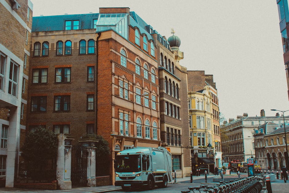 A street scene featuring a blue and white waste collection vehicle parked in front of a multi-storey red brick building with distinctive arched windows and a modern teal roof. To the right, historic architecture with ornate facades and a rounded turret topped with a dome are visible, indicating a mixture of building styles typical of urban areas. The vehicle is positioned adjacent to an entrance gate with stone pillars, and nearby, several bollards and bicycles are lined up along the pavement. The surrounding environment includes other buildings with classic and contemporary architectural elements, a lamppost, and a busy street atmosphere with vehicles and pedestrians further in the background. The scene is brightly lit, capturing the details of the materials, textures, and colors, highlighting the context of urban rubbish collection and private waste removal services managed by Rubbish Clearance Stockwell, serving the general waste management needs of the area.