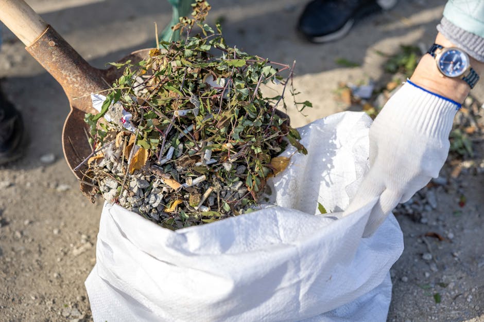 A person wearing a white glove is holding a large white woven bag filled with miscellaneous garden waste, including leafy branches, small twigs, and bits of soil-covered debris. The bag appears to be placed on a gravel surface, and a rusted shovelful of soil and plant matter is being held above the bag, ready for transfer. In the background, there are partial views of a person wearing a watch and black shoes, along with blurred outdoor surroundings. The scene suggests a site for collection or removal of garden or yard waste, possibly as part of an independent rubbish clearance service. The natural lighting highlights the textures of the leaves, twigs, and gravel, creating a clear, detailed image suitable for illustrating waste removal or rubbish clearance activities typically handled by companies such as Rubbish Clearance Stockwell.