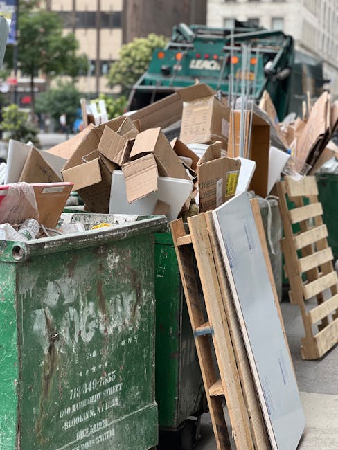 A large green waste disposal bin filled with a mix of flattened cardboard boxes, some small and others larger, as well as miscellaneous packaging materials. Several of the boxes are open and partially crushed, revealing their corrugated cardboard texture with brown and white surfaces. Leaning against the bin is a wooden pallet with a partially visible sheet of glass or plastic resting on it. Behind the bin, a street scene is visible with a green waste collection truck parked nearby, and blurred background buildings and trees. The scene appears to be outdoors on a city street, with natural light illuminating the rubbish and surroundings, aligning with independent rubbish clearance services that handle on-site waste removal and disposal, such as those offered by Rubbish Clearance Stockwell.