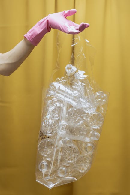 A person wearing a white glove is holding a large white woven bag filled with miscellaneous garden waste, including leafy branches, small twigs, and bits of soil-covered debris. The bag appears to be placed on a gravel surface, and a rusted shovelful of soil and plant matter is being held above the bag, ready for transfer. In the background, there are partial views of a person wearing a watch and black shoes, along with blurred outdoor surroundings. The scene suggests a site for collection or removal of garden or yard waste, possibly as part of an independent rubbish clearance service. The natural lighting highlights the textures of the leaves, twigs, and gravel, creating a clear, detailed image suitable for illustrating waste removal or rubbish clearance activities typically handled by companies such as Rubbish Clearance Stockwell.