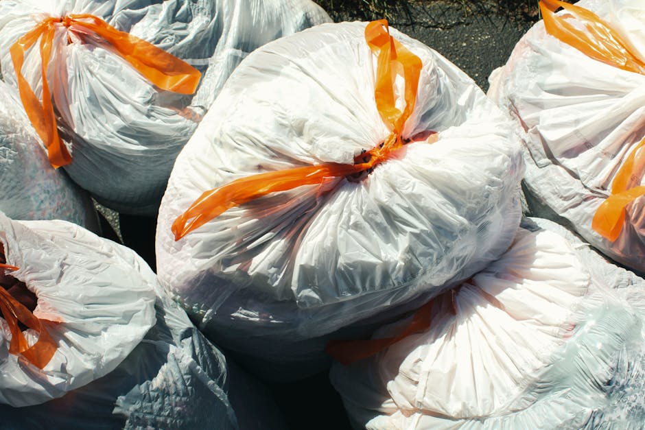 A group of white plastic rubbish bags filled with waste, tightly knotted at the top with orange ties, resting on a paved outdoor surface. The bags are large, slightly crinkled, and appear to contain household waste or discarded materials, with some showing faint folds and creases in the plastic. The bags are positioned closely together, with one in the center prominently visible and others surrounding it, indicating an outdoor rubbish collection or clearance scene. The sunlight creates subtle reflections on the plastic surfaces, highlighting the texture and the semi-translucent quality of the bags. This scene exemplifies a typical site for private waste disposal or alternative rubbish removal services, such as those provided by Rubbish Clearance Stockwell, which often handle on-site rubbish collection and environmentally responsible disposal, especially in urban or residential settings. The environment suggests an outdoor area, possibly a driveway or a pavement, prepared for collection or transfer to waste removal vehicles, fitting within the context of professional rubbish clearance and waste management efforts.