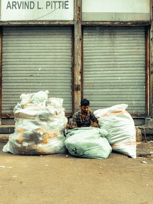 A man with dark hair and a beard, wearing a patterned shirt, is sitting on the ground between four large trash bags filled with various waste materials. The bags are made of plastic, with two being transparent and showing mixed rubbish inside, while the other two are opaque, one white and the other light green. The scene is set outdoors on a dirt or concrete surface in front of a weathered, closed metal roller-shutter door with visible rust and discoloration, mounted on a building with a partially visible sign reading 'ARVIND L. PITTIE' at the top left. The environment appears urban and somewhat neglected, with the focus on waste collection and disposal, reflecting a private or independent rubbish removal activity. The lighting suggests daytime with natural light casting soft shadows, emphasizing the textures of the plastic bags and the metal door behind.